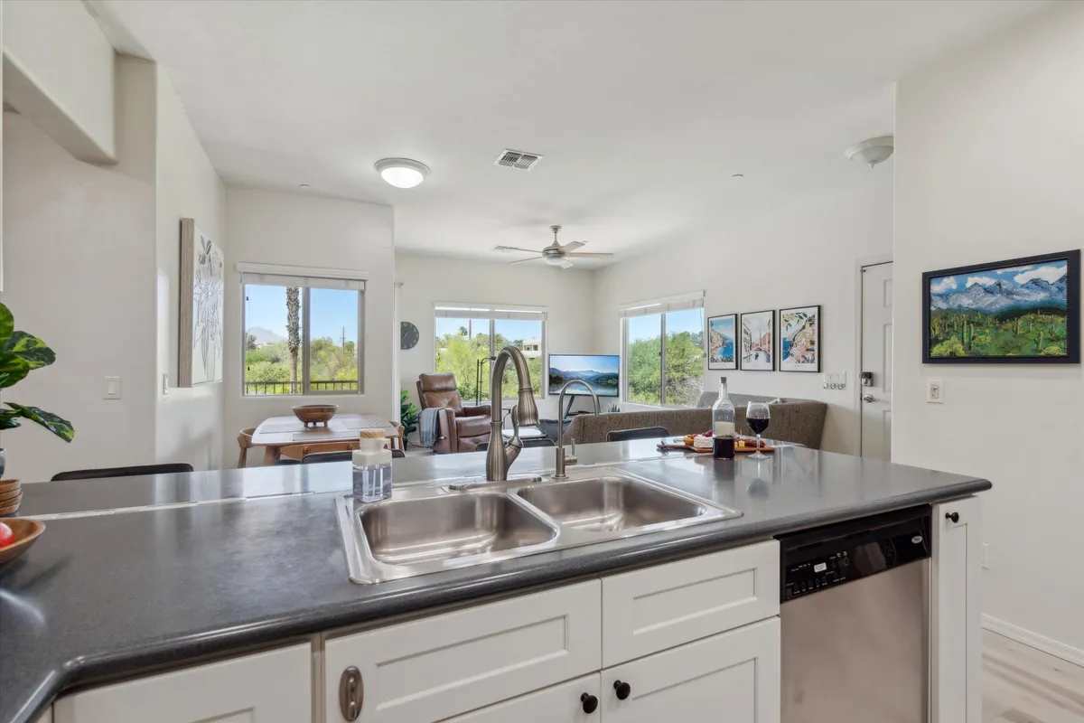 A bright, welcoming space that blends a kitchen island (with sink), a dining table set for four, and a visible living area with comfortable seating and TV. Large windows usher in natural light, and the open floor plan ensures easy interaction between spaces.