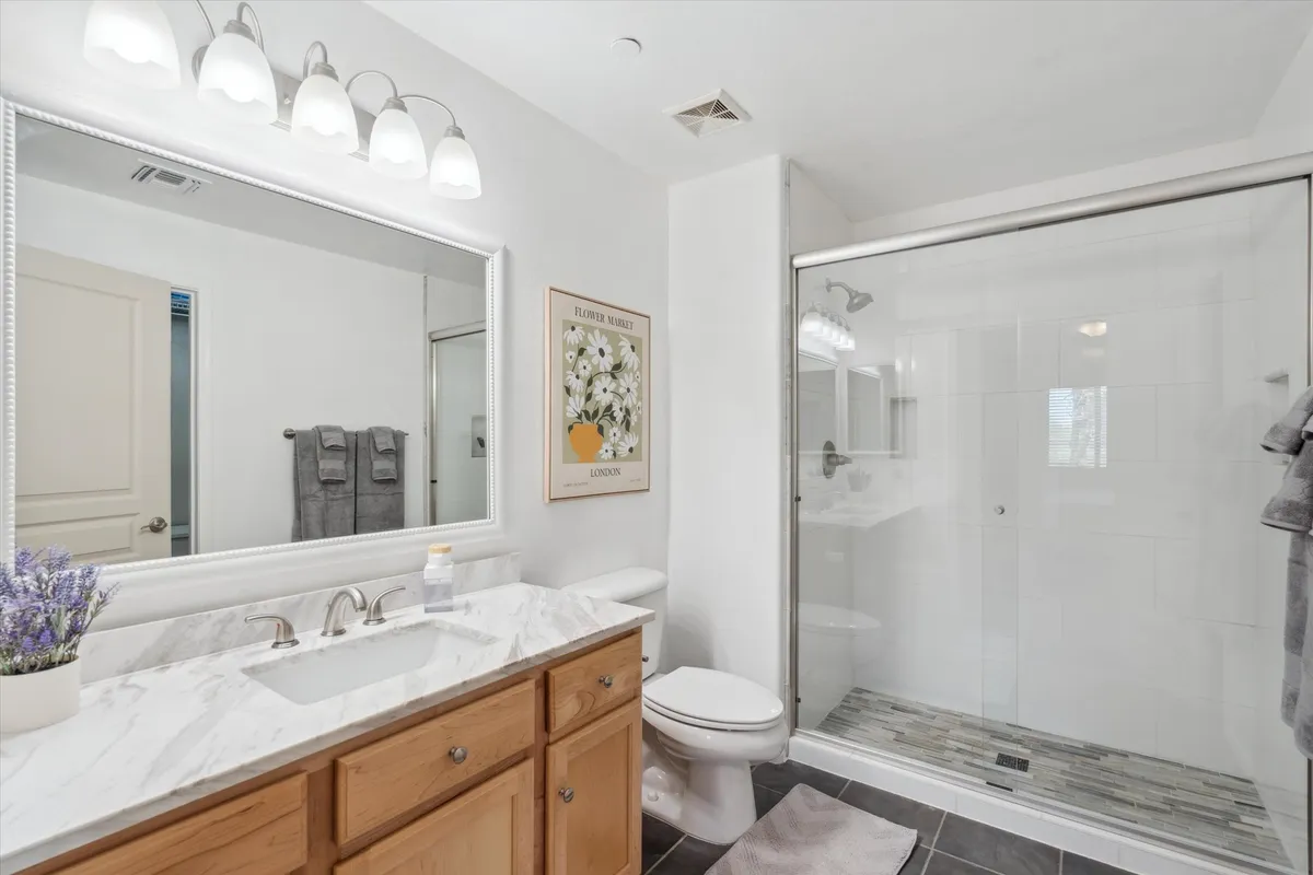 A bright, modern bathroom featuring a double-sink vanity, large mirror, walk-in shower enclosure, and flower-themed artwork. The space is well-lit, emphasizing cleanliness and a spa-inspired environment perfect for morning rituals or evening relaxation.