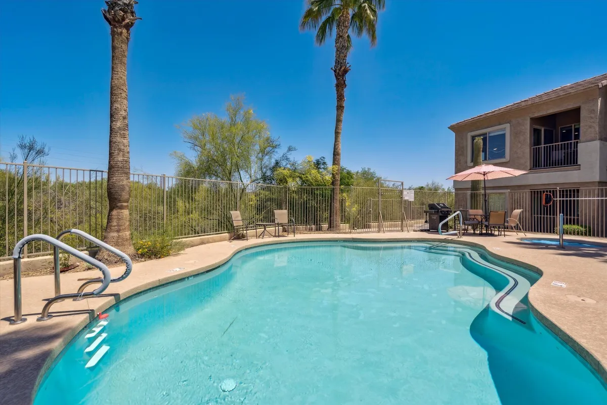 This exterior shot captures a sparkling swimming pool surrounded by palm trees and an inviting outdoor seating area. The condo’s balcony is visible, hinting at the proximity of the residence to the resort-style amenities. The scene is alive with activities like swimming, sunbathing, and socializing, all framed by beautiful landscaping and clear skies.