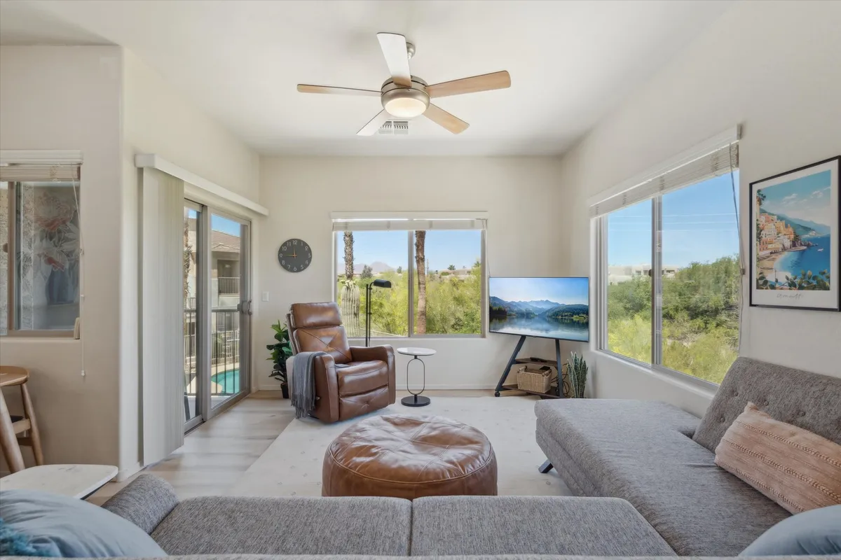 A bright, open living area featuring large windows that allow natural light to pour in. The comfortable seating area is oriented towards a TV displaying a scenic landscape, and decorative artwork adds character. The windows frame views of the outdoors, hinting at mountain or park vistas beyond.