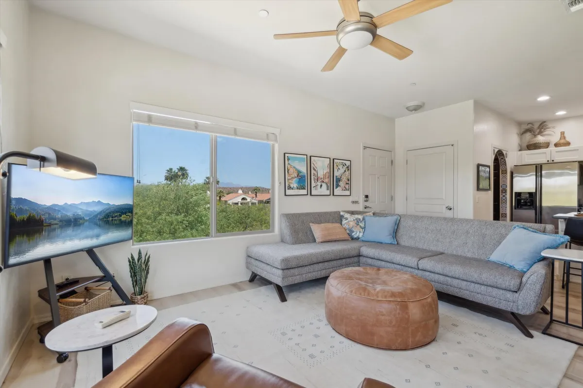 A spacious living room with a large sectional sofa perfectly positioned to enjoy both a mounted television and the view through a sunlit window. Artwork adds a stylish touch, and the window offers a glimpse of the outdoors, bringing in natural light.