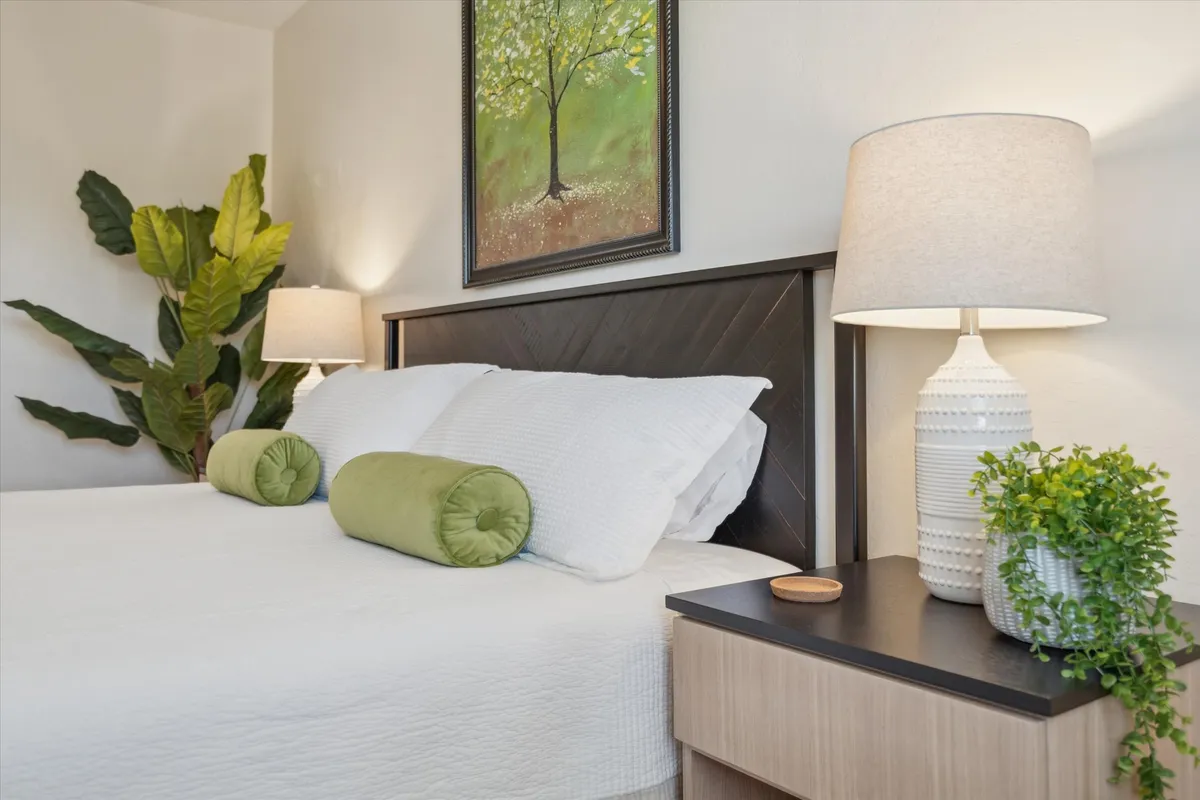 A serene bedroom showcasing a king-sized bed with inviting white bedding and green accent pillows. Framed artwork of a tree hangs above the bed, complemented by soft-light table lamps and an indoor plant, creating a calm and cozy retreat suited for restful nights.