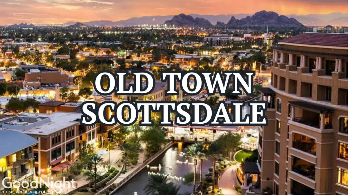 A twilight view over Old Town Scottsdale, featuring illuminated signage, waterway reflections, and surrounding mountains. People can be seen enjoying evening strolls, dining, and live entertainment along vibrant, well-lit streets.