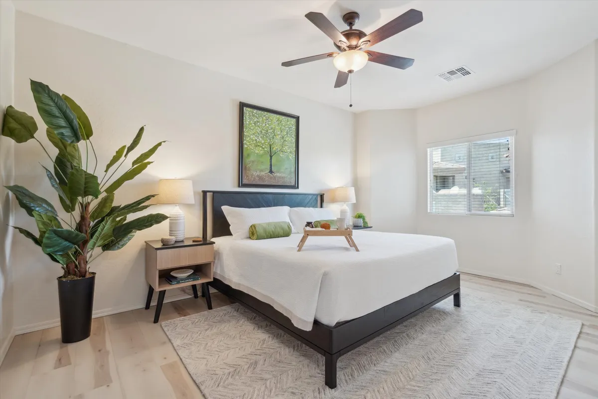 A tranquil bedroom scene featuring a king-sized bed dressed in crisp white linens, accented by a green decorative roll pillow. The nightstand holds a lamp for soft lighting, while a framed painting of a tree and an indoor plant add touches of nature and serenity. Sunlight filters through a nearby window, enhancing the room's restful ambiance.