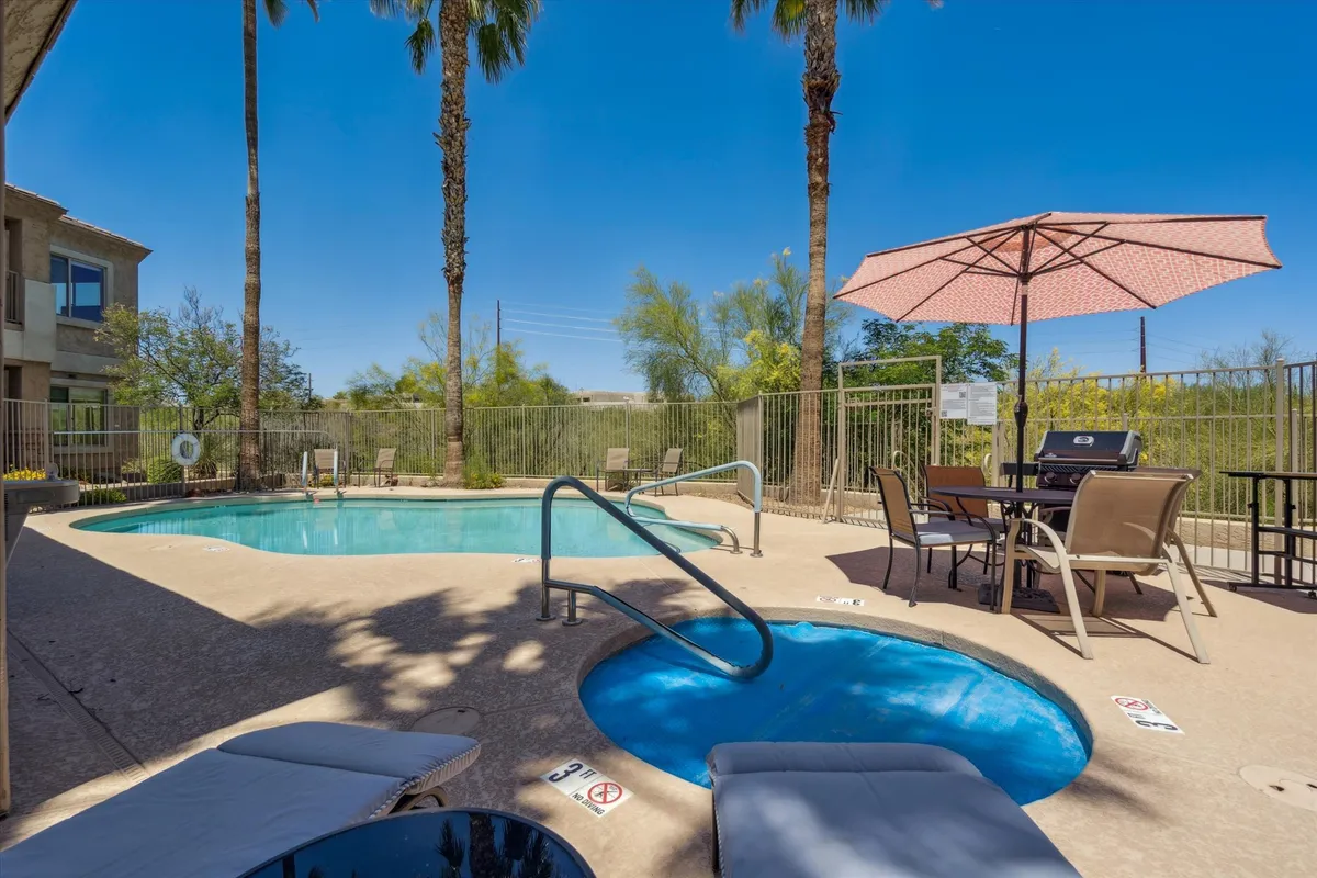 An exterior scene featuring a sparkling swimming pool surrounded by palm trees, a bubbling hot tub, outdoor dining area, and shaded umbrellas. The setting is lively yet serene, perfectly blending recreation and relaxation. The pool and hot tub are at the heart of the image, inviting residents to unwind or socialize.